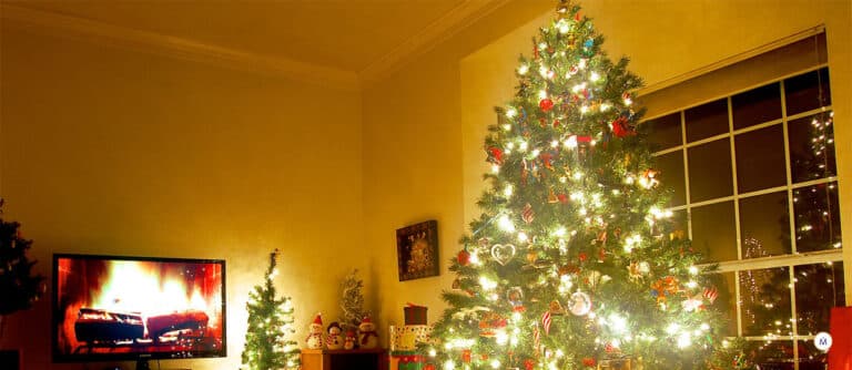 Un árbol de Navidad decorado con muchas luces y bolas, junto a una televisión con imágenes de una chimenea